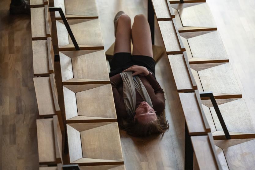 Das Bild zeigt eine Performerin, die auf dem Rücken zwischen zwei Reihen schlichter, geometrisch geformter Holzbänke in einer Kirche liegt. Ihre Beine sind leicht angewinkelt, sie trägt ein braunes Oberteil, schwarze Shorts und ein Tuch um die Schultern. Ihr Blick ist nach oben gerichtet, ihr Gesicht wirkt ruhig und kontemplativ.