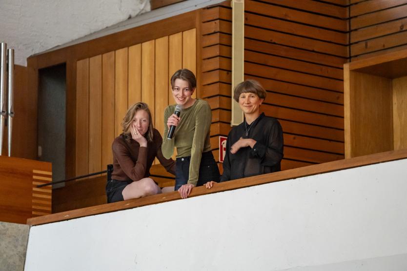 Drei Personen stehen auf einer Empore in einem holzverkleideten Raum. Eine von ihnen hält ein Mikrofon und spricht, die anderen blicken ruhig in Richtung Kamera.