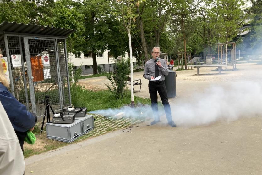 Während eine Nebelmaschine im Vordergrund aktiv ist, spricht ein Mann mit Mikrofon und Notizen vor einer kleinen Gruppe im Park.