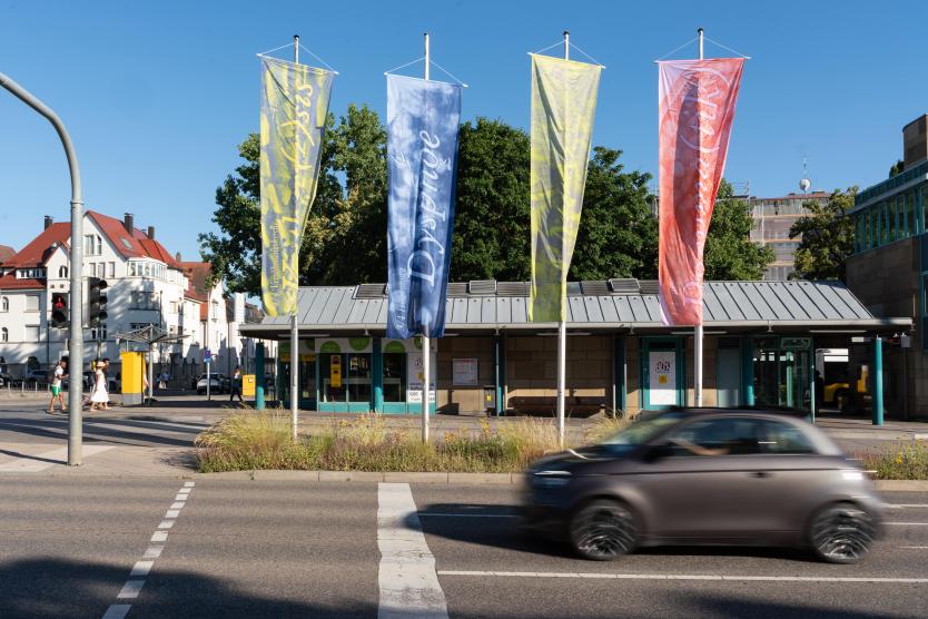 Zu sehen sind 4 Fahnen, zwei gelbe, eine blaue und eine rote Fahne die sich an einer befahrenen Straße befinden. Im Hintergrund ist eine Bushaltestelle zu sehen.