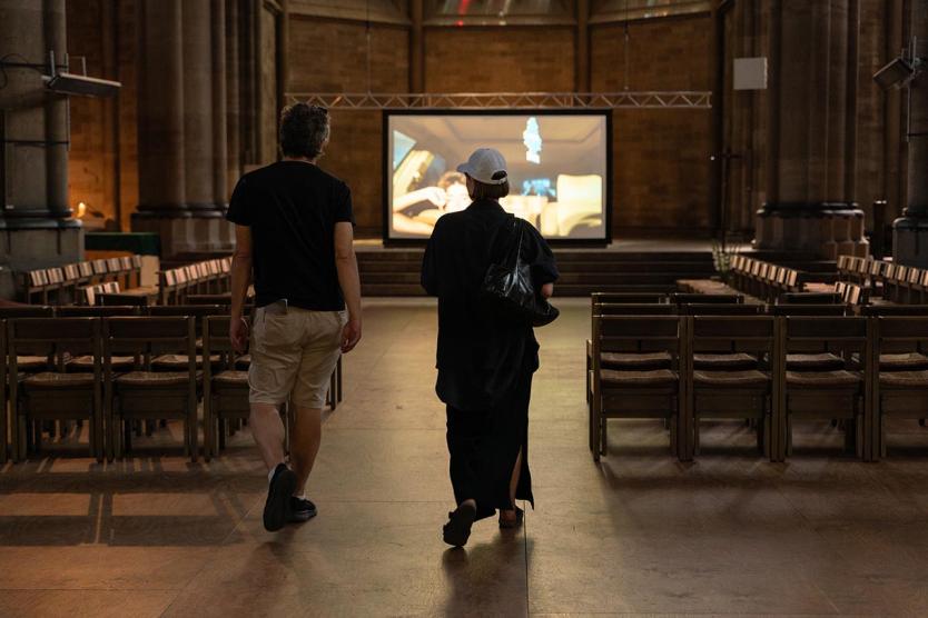 Zwei Personen betreten einen großen, ruhigen Kirchraum, in dem ein Video auf einer Leinwand gezeigt wird.