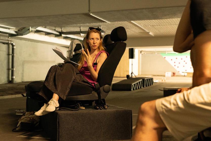 Eine Person sitzt auf einem Autositz in einer Tiefgarage, spricht engagiert und gestikuliert, ein Blatt Papier in der Hand. 