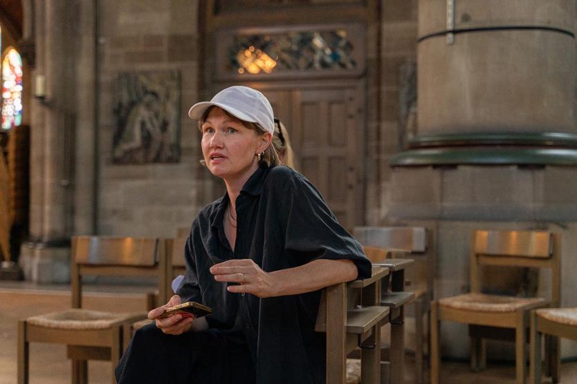 Person mit weißer Kappe spricht im Kirchenraum, umgeben von Holzstühlen und historischer Architektur.