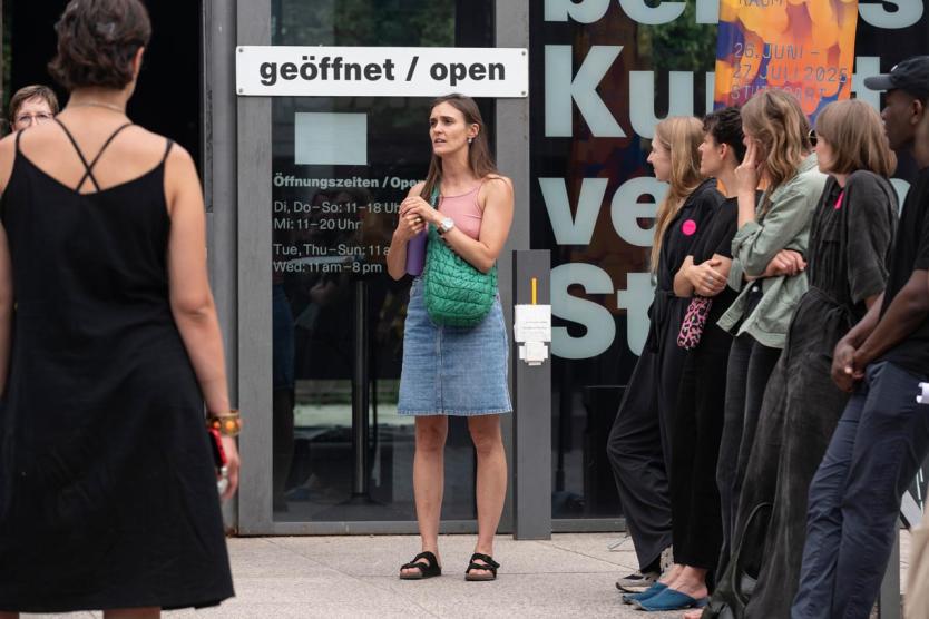 Person spricht vor einer Gruppe vor dem Eingang eines Kunstinstituts, Schild „geöffnet / open“ im Hintergrund.