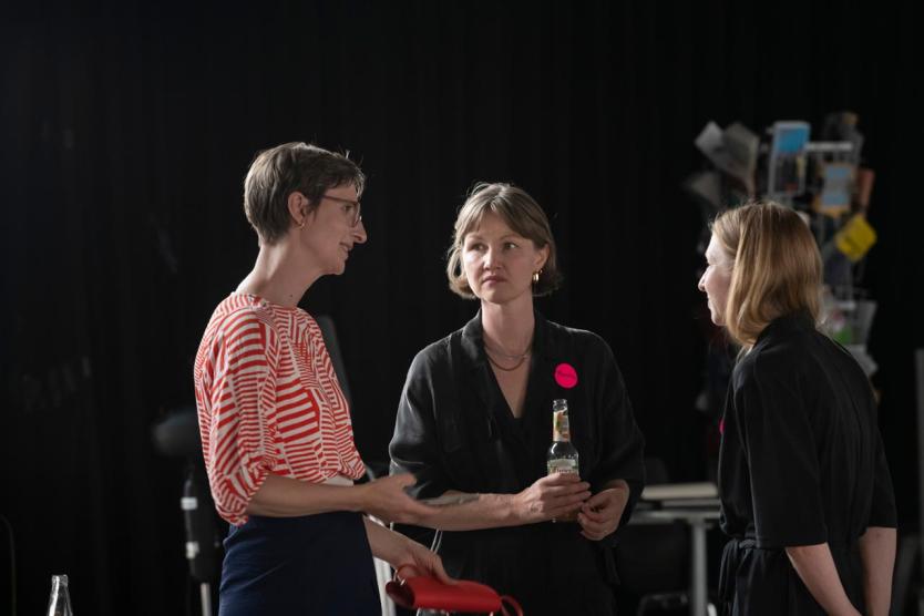 Drei Personen im Gespräch vor schwarzem Hintergrund, mittig eine Getränkeflasche in der Hand.