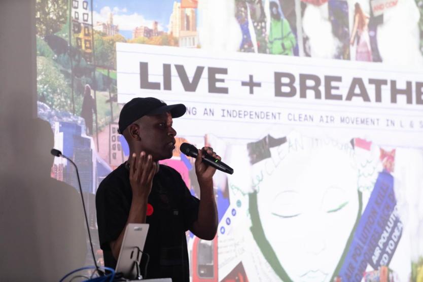 Person mit Mikrofon vor farbiger Präsentationsfolie mit der Aufschrift „LIVE + BREATHE“.