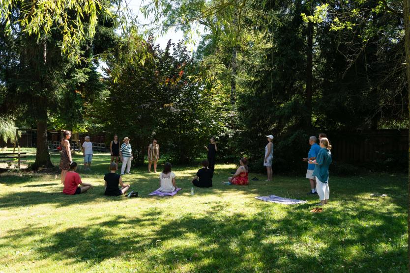 Eine Gruppe an Personen sitzen und stehen auf einer Wiese, umgebend von Bäumen, und eine Personen leitet eine Entspannungsübung an.