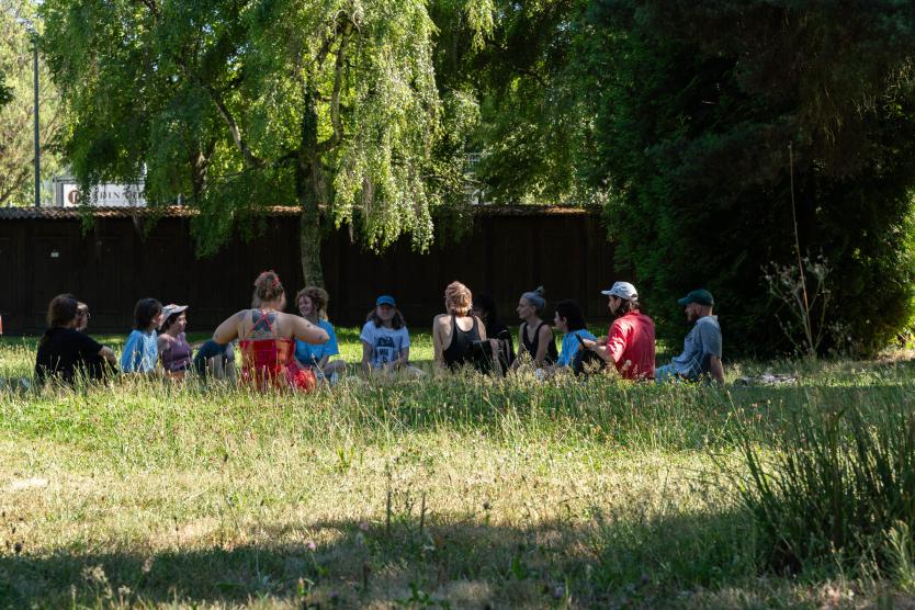 Eine Gruppe an Personen sitzen auf einer Wiese, umgebend von Bäumen, sie sind in einem Gespräch.