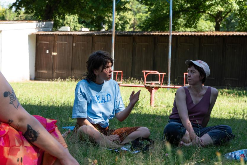 Drei Personen sitzen im Gras und unterhalten sich. Die eine Person gestikuliert mit ihrer Hand. Im Hintergrund sind hölzerne Kabinen zu sehen.