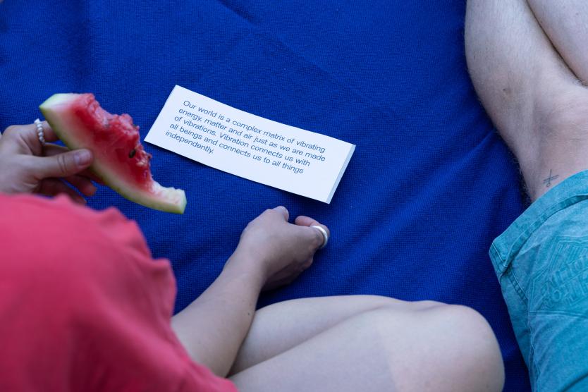 Die Szene zeigt eine Person die auf einer blauen Picknickdecke eine Wassermelone isst und einen Text auf einem kleinen Zettel liest. 