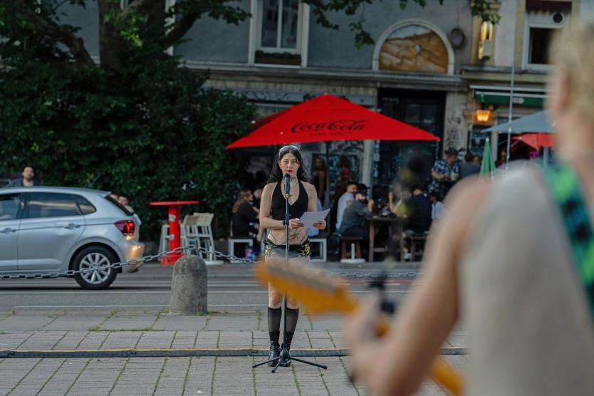 Das Bild zeigt eine Performerin, die mitten auf einer Straße vor einem roten Sonnenschirm steht und mit festem Blick einen Text in ein Mikrofon spricht. Im Vordergrund ist unscharf eine Person mit Gitarre zu sehen.
