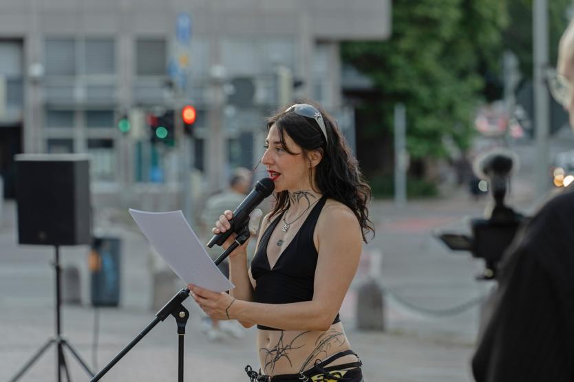Das Bild zeigt eine Frau mit langen dunklen Haaren, die in ein Mikrofon spricht und dabei ein Blatt Papier in der Hand hält. Sie trägt ein schwarzes Oberteil, Sonnenbrille auf dem Kopf und ist stark tätowiert.
