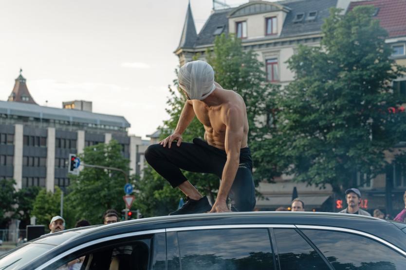Das Bild zeigt eine Person mit nacktem Oberkörper und Bandagen um den Kopf. Die Person kniet auf dem Dach eines schwarzen Autos, umgeben von Stadtgebäuden und Bäumen. Einige Menschen schauen von unten zu.