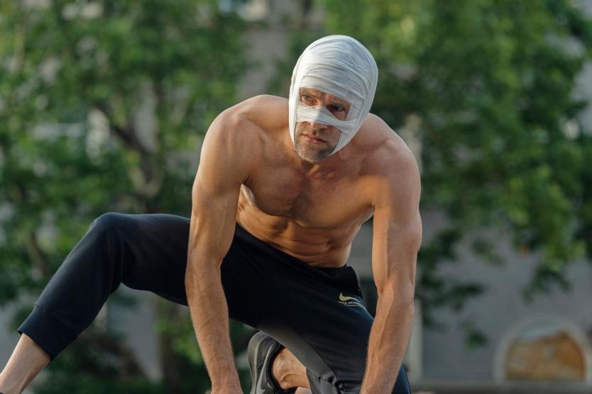 Das Bild zeigt eine Person mit nacktem Oberkörper und Bandagen um den Kopf. Der Körper ist angespannt, der Blick ernst. Im Hintergrund sind Bäume und verschwommene Gebäudeteile zu sehen.