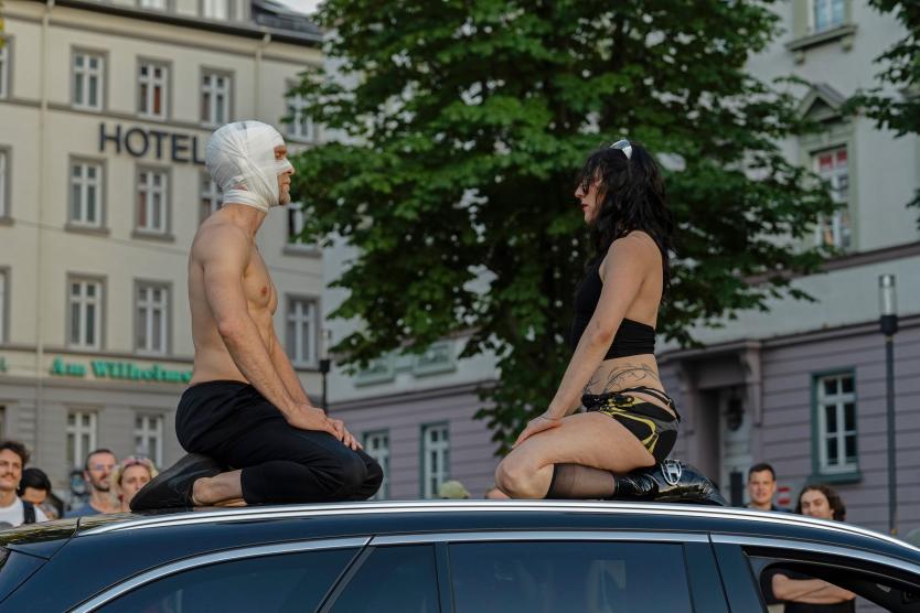Zwei Performer*innen knien sich gegenüber auf dem Dach eines Autos und schauen sich intensiv an. Um sie herum beobachten viele Menschen das Geschehen.
