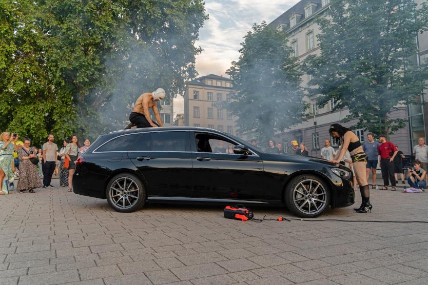 Das Bild zeigt eine Performance im Stadtraum. Eine Person kniet mit nacktem Oberkörper und Maske auf dem Dach eines schwarzen Autos, während eine andere Person vorne an der Motorhaube steht. Rauch steigt auf und viele Menschen schauen zu.