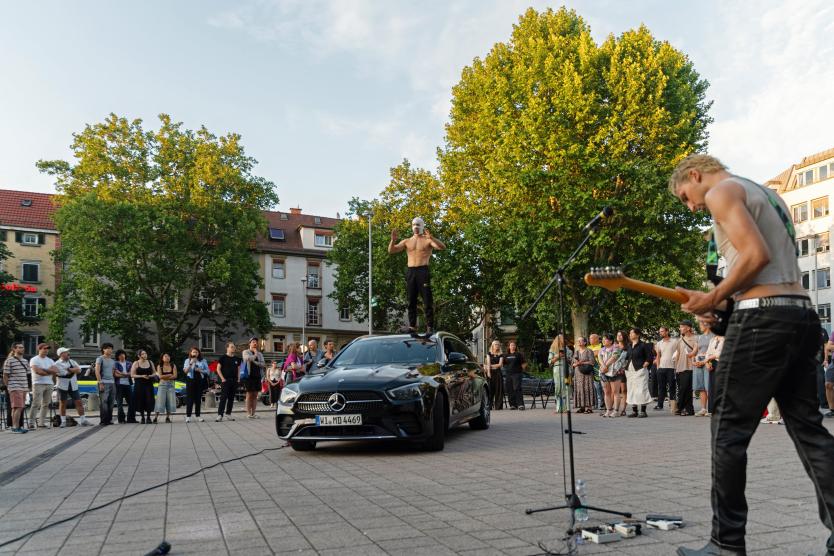 Das Bild zeigt eine Performance im Freien. Eine Person steht mit nacktem Oberkörper und Maske auf dem Dach eines schwarzen Autos, während eine andere Gitarre spielt. Viele Menschen stehen im Kreis und schauen zu.