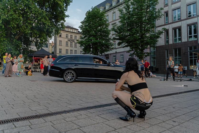 Das Bild zeigt ein schwarzes Auto auf einem Platz in der Stadt. Eine Person in schwarzer Kleidung hockt davor, während weiter hinten jemand Gitarre spielt und singt. Menschen stehen oder sitzen rundherum und schauen zu.