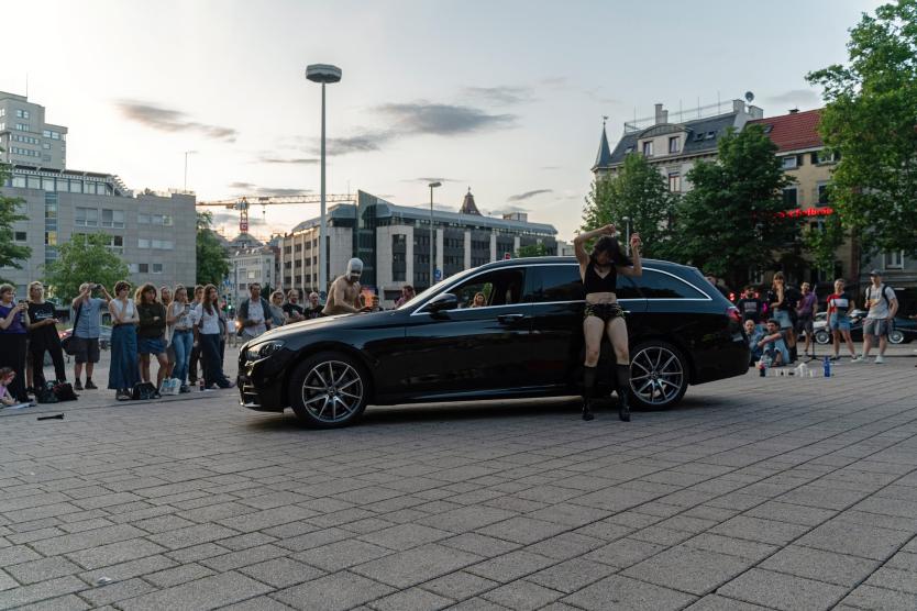 Eine Performance auf einem öffentlichen Platz. Zwei Performer*innen bewegen sich kraftvoll und expressiv um ein schwarzes Auto. Eine Person tanzt mit Mikrofon, eine andere steht auf dem Kofferraum. Das Publikum bildet einen Halbkreis um die Szene.