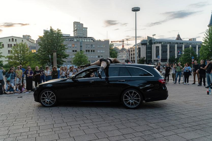 Auf einem öffentlichen Platz liegt eine Performerin auf einem fahrenden schwarzen Auto, halb ausgestreckt über Dach und Fenster. Zahlreiche Zuschauerinnen und Zuschauer stehen im Halbkreis und beobachten die Szene aufmerksam.