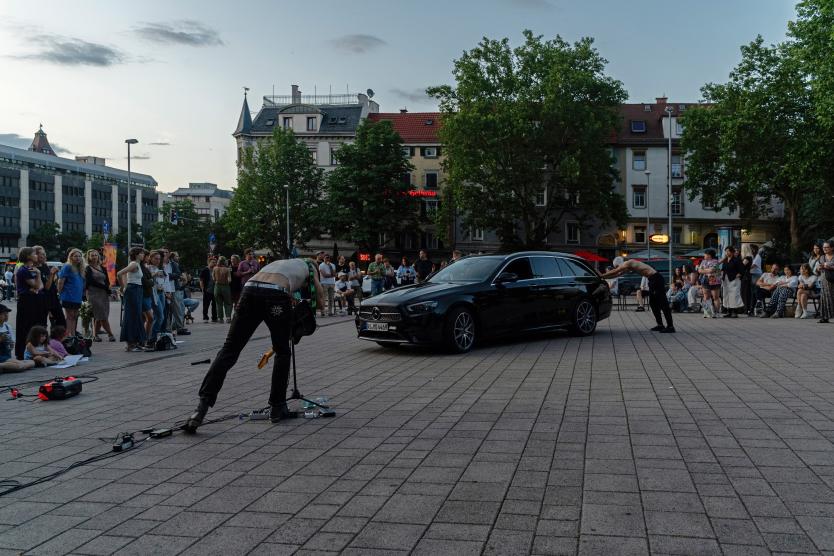 Eine Performance auf einem öffentlichen Platz. Zwei Personen bewegen sich dynamisch um ein schwarzes Auto. Eine spielt E-Gitarre, die andere beugt sich nach vorn. Viele Menschen stehen oder sitzen im Kreis und schauen zu.