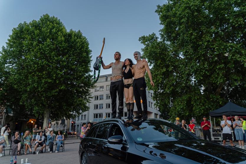 Drei Performer*innen stehen auf dem Dach eines schwarzen Autos und nehmen eine Pose ein. Eine Person hält eine Gitarre in die Höhe. Um das Auto herum sitzen und stehen Zuschauende, viele applaudieren.