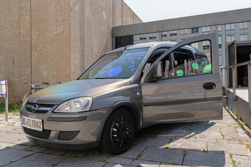 Ein silbernes Auto, blaue Punkte spiegeln sich in Fenstern, steht vor einem Gebäude und die Vordertür ist geöffnet. In dem Auto sitzt eine Person.