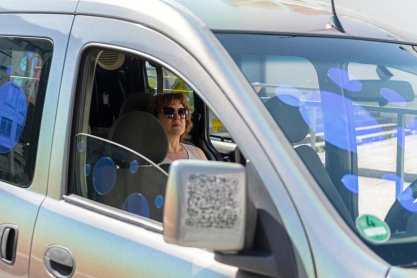 Frau mit Sonnenbrille sitzt in silbernem Auto, blaue Punkte spiegeln sich in Fenstern.