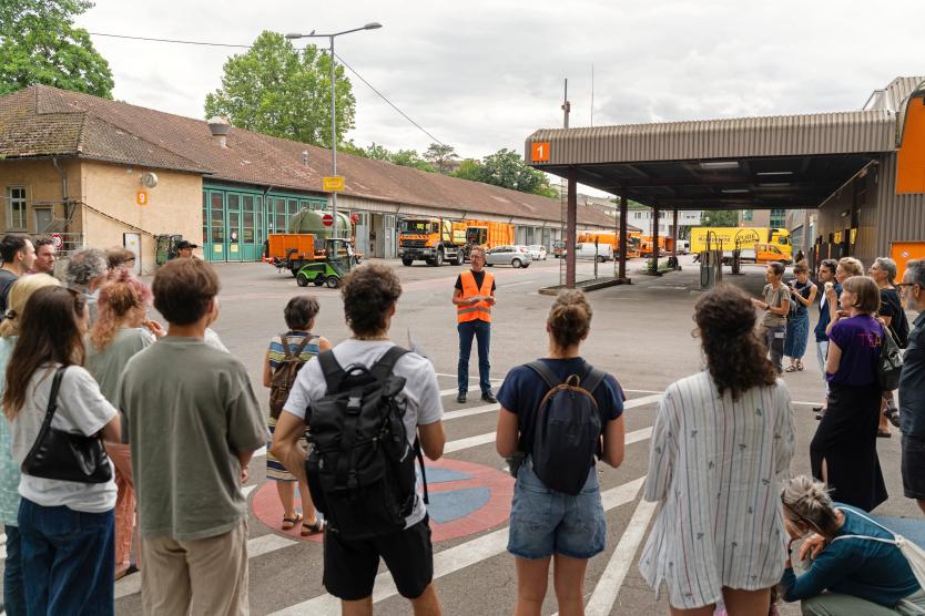 Eine Gruppe von Besucher*innen steht auf dem Gelände der Abfallwirtschaft und hört einer Person mit orangefarbener Warnweste zu. Im Hintergrund sind Einsatzfahrzeuge, Hallen und Container zu sehen. 