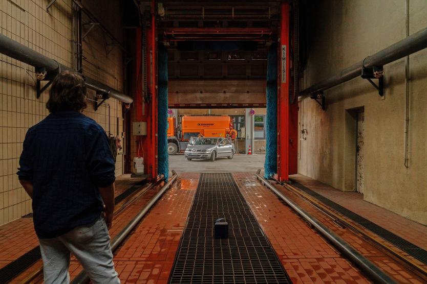 Eine Person steht in einer leeren Waschanlage und blickt nach draußen, wo ein silbernes Auto vor einem großen orangefarbenen Fahrzeug geparkt ist.