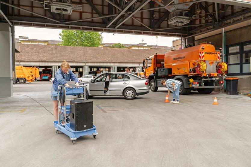 Auf dem Gelände der Abfallwirtschaft findet eine Performance statt. Drei Personen bewegen sich zwischen einem silbernen Auto, orangefarbenen Einsatzfahrzeugen und einem blauen Technikwagen. Eine Person arbeitet am Lautsprecher, eine andere greift nach einem Verkehrshütchen.