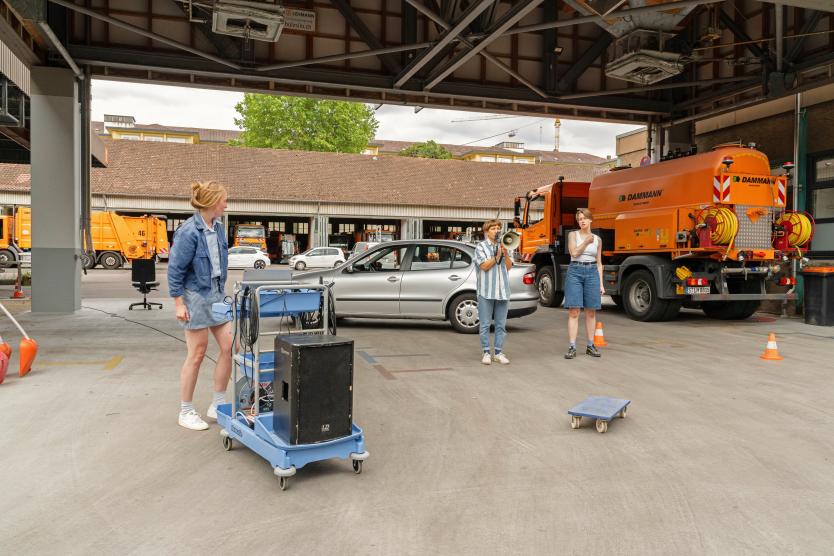 Auf dem Gelände der Abfallwirtschaft stehen drei Personen zwischen orangefarbenen Einsatzfahrzeugen und einem silbernen Auto. Eine Person spricht durch ein Megafon, eine andere steht an einem blauen Rollwagen mit Lautsprecher. 