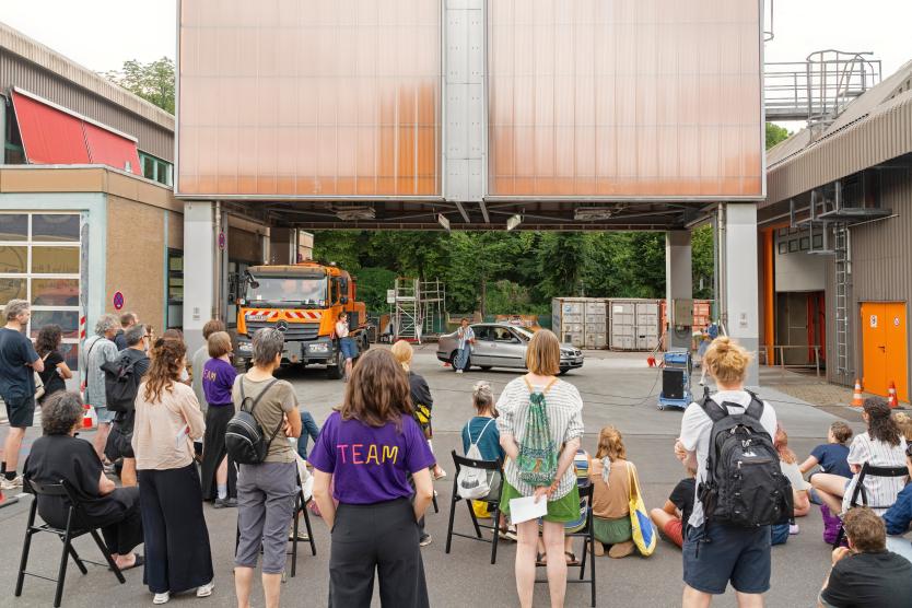 Eine Gruppe von Menschen steht und sitzt auf dem Gelände der Abfallwirtschaft und schaut auf eine Performance. Vor einer orangefarbenen Überdachung stehen ein silbernes Auto und ein städtisches Nutzfahrzeug. Dazwischen bewegen sich Performer*innen.