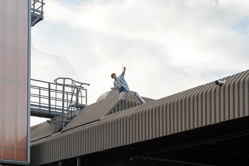 Eine Person steht auf dem Dach eines Industriegebäudes auf dem Gelände der Abfallwirtschaft. Mit ausgestrecktem Arm wird eine bewegte Geste gezeigt – Teil einer Performance in großer Höhe vor dem Himmel.