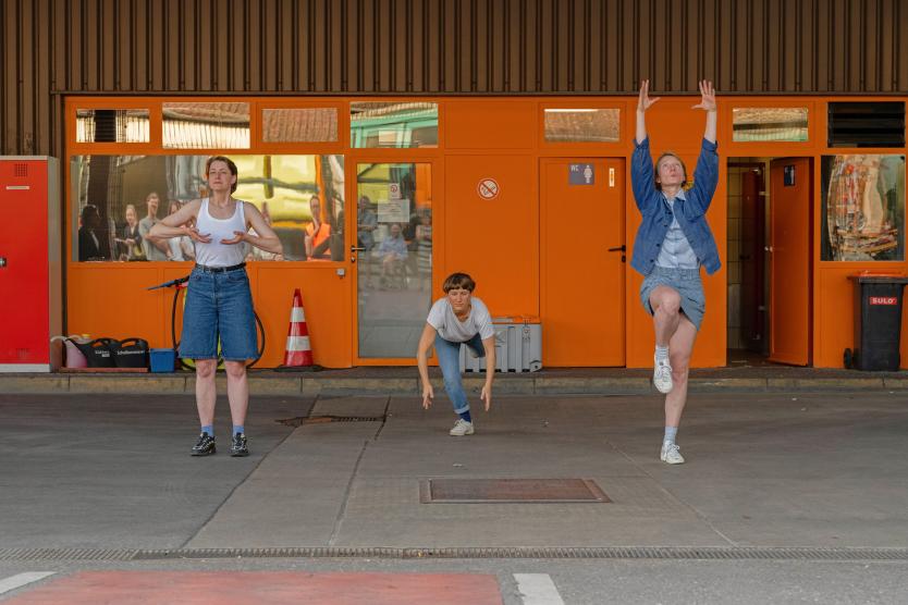 Drei Personen stehen auf dem Gelände der Abfallwirtschaft vor einer großen orangefarbenen Wand. Jede spricht in ein Mikrofon und hält Papier in der Hand. Ein blauer Rollwagen mit Technik und ein Lautsprecher stehen davor. Die Szene zeigt eine Performance im Freien.