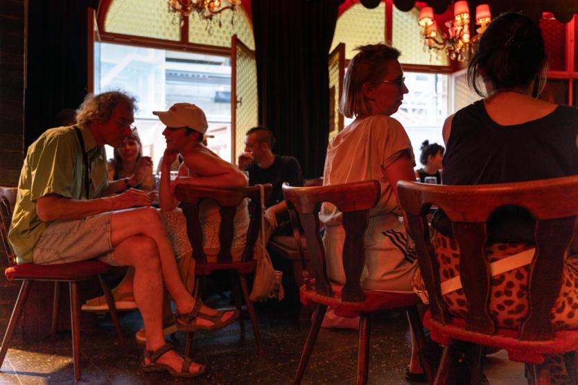 Ein stimmungsvoll beleuchteter Raum mit rotem Licht. Menschen sitzen in kleinen Gruppen auf Holzstühlen, sprechen miteinander, einige lehnen sich einander zu. Fenster sind geöffnet, draußen ist Tageslicht zu sehen.