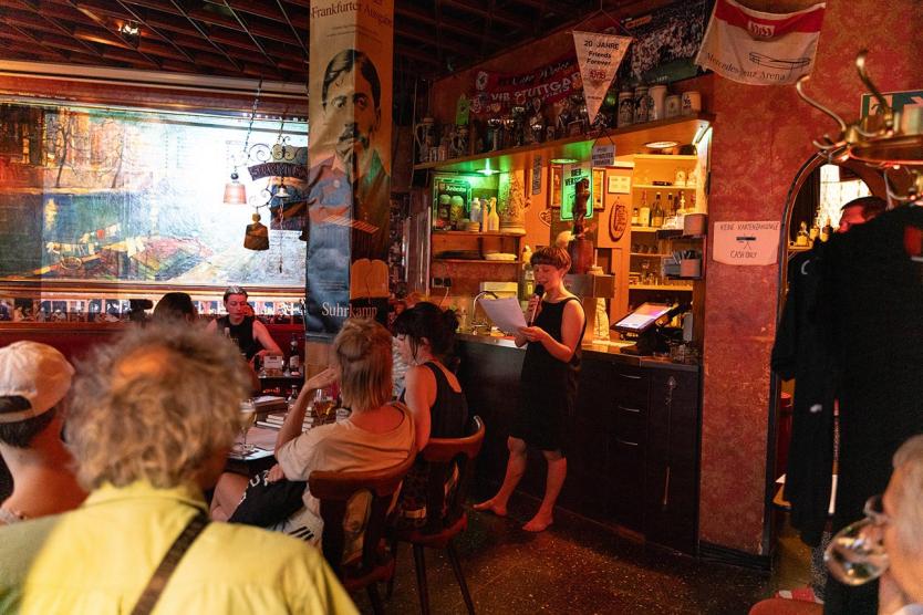 Ein gemütlicher, rot-gold beleuchteter Barraum. Menschen sitzen an Tischen, hören einer barfüßigen Person zu, die mit Mikrofon vorliest. Die Atmosphäre wirkt aufmerksam und entspannt.