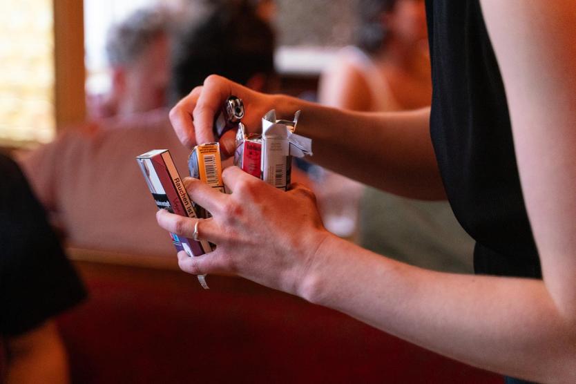 Eine Person hält mehrere bunte Zigarettenschachteln und ein Feuerzeug in der Hand. Im Hintergrund sitzen unscharf weitere Menschen in gemütlicher Bar-Atmosphäre.