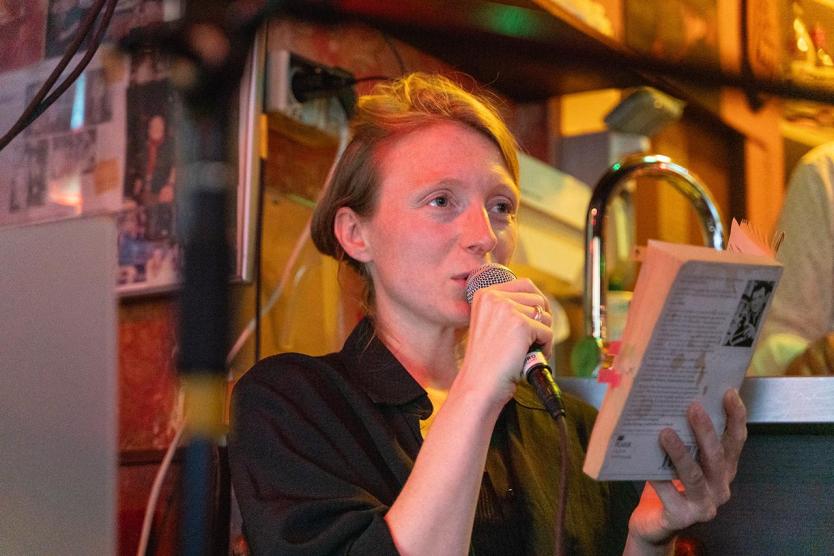Eine Frau mit offenem Haar liest in einer Bar aus einem Buch vor. Sie spricht ins Mikrofon und hält das Buch mit Markierungen in der anderen Hand. Die Atmosphäre ist intim und konzentriert.