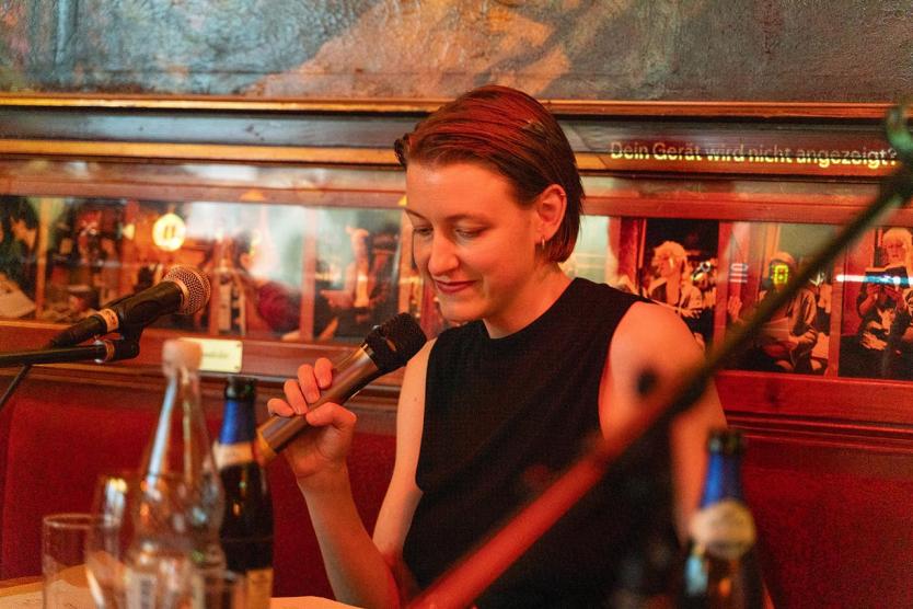 Szene in Bar-Atmosphäre: Person mit Mikrofon beim Sprechen oder Vorlesen, vor sich Getränke, Mikrofone und Notizen.