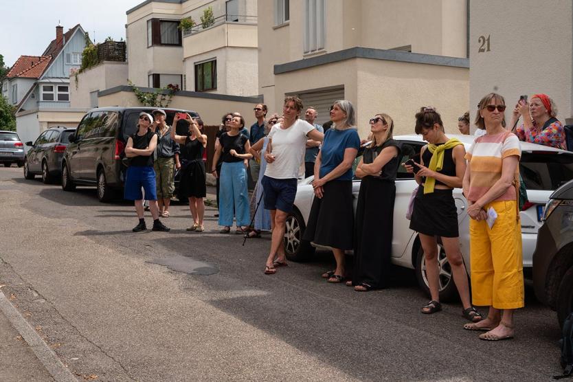 Eine Gruppe von Menschen steht auf einer Wohnstraße vor hellen Gebäuden. Einige blicken aufmerksam nach oben, andere filmen oder fotografieren mit ihren Handys. Autos sind entlang des Gehwegs geparkt, im Hintergrund Häuser mit Balkonen und gepflegten Fassaden. Die Szene wirkt wie ein gemeinschaftliches Zuschauen bei einer Aktion im öffentlichen Raum.