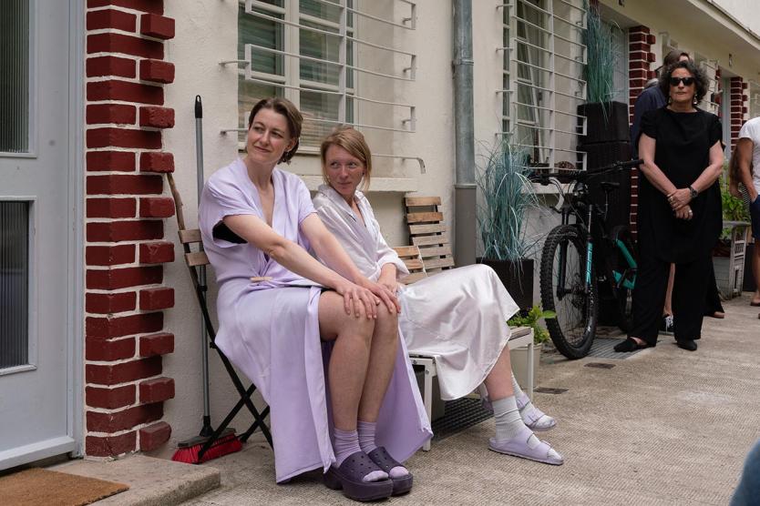 Zwei Personen in hellen Gewändern sitzen vor einem Wohnhaus. Weitere Menschen stehen im Hintergrund. Ein Fahrrad lehnt an der Wand.