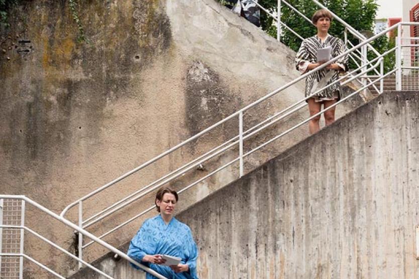 Drei Personen stehen auf unterschiedlichen Abschnitten einer Treppe zwischen zwei Wohngebäuden. Die Szenerie wirkt inszeniert: Alle tragen auffällige, gemusterte Gewänder und halten jeweils ein Blatt Papier in den Händen. Die Architektur im Hintergrund zeigt helle Fassaden mit roten Fensterläden.