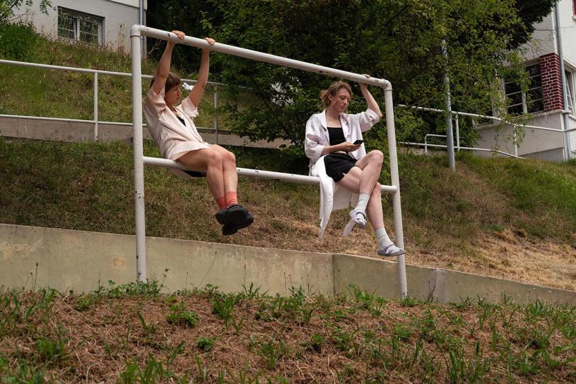 Zwei Personen sitzen nebeneinander auf einem weißen Geländer, das parallel zu einem Hang verläuft. Eine Person hängt mit beiden Armen an der oberen Querstange und blickt konzentriert zur Seite, die Beine angewinkelt über der unteren Strebe. Die andere Person sitzt entspannt mit überschlagenen Beinen, eine Hand am Geländer, die andere hält ein Smartphone. Im Hintergrund: Hang mit Wiese, Sträuchern und Wohngebäuden mit roten Fensterläden.