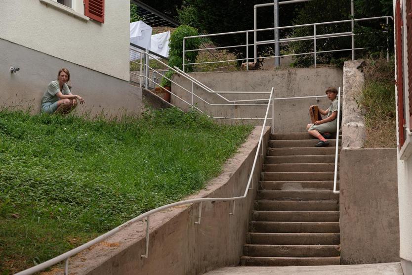 Zwei Personen in heller Kleidung sitzen sich über eine Treppe hinweg gegenüber. Wohngebäude und Grünflächen umgeben die Szene.