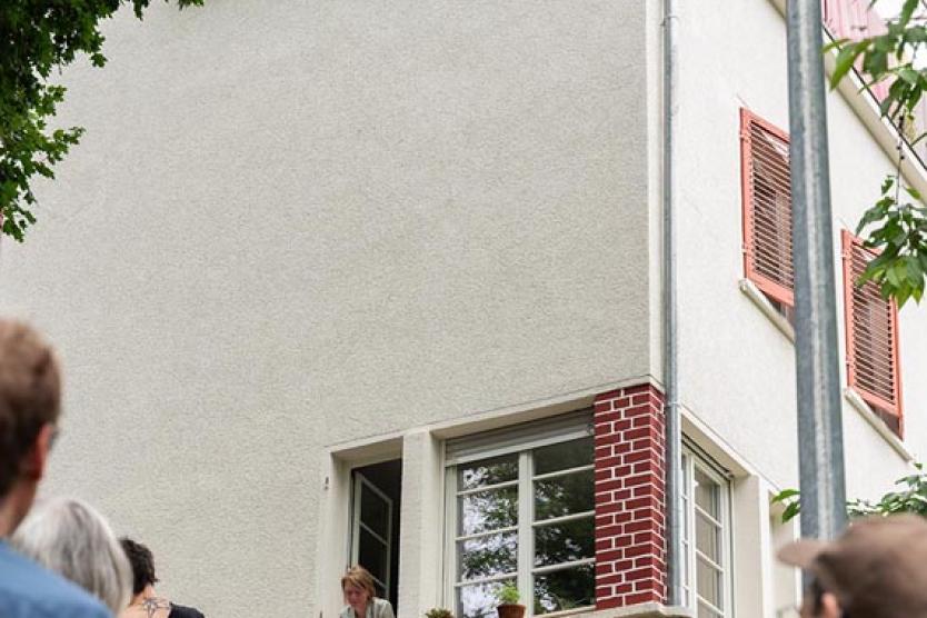 Eine Gruppe von Menschen steht vor einem Wohnhaus mit roten Fensterläden, zwei Personen schauen aus geöffneten Fenstern im Erdgeschoss und Obergeschoss auf die Szene herab.