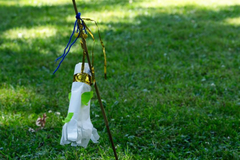 Das Bild zeigt einen Stock, der in einer Wiese steckt und an dem ein handgemachtes Windspiel hängt.