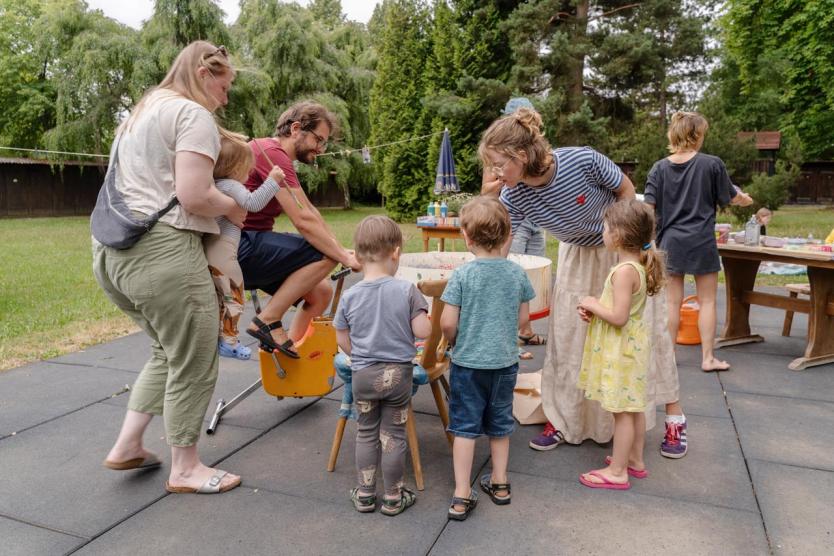 Mehrere Kinder und Erwachsene versammeln sich um ein buntes Mitmachgerät im Freien, ein Mann tritt in die Pedale eines Fahrrads, während um ihn herum gemalt, beobachtet und gelacht wird.