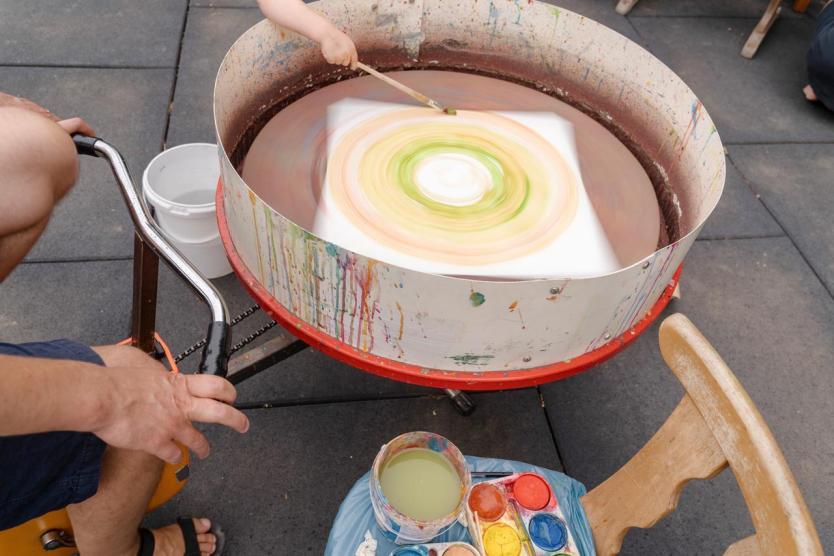 Ein großes rotierendes Farbrad wird mit einem Pinsel bemalt, während daneben ein Pedal angetrieben wird; auf dem Tisch davor stehen Wasserbecher und bunte Farbtöpfe – kreative Bewegung trifft auf Farbe.