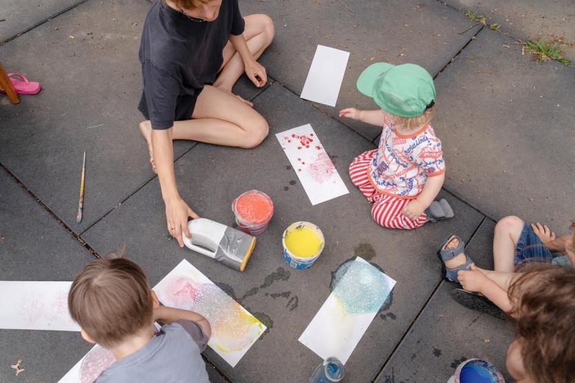 Kinder und eine erwachsene Person sitzen im Kreis auf Steinplatten, rundherum bunte Farben in Bechern und Papierbögen mit Seifenblasenmustern – gemeinsames kreatives Arbeiten im Freien.